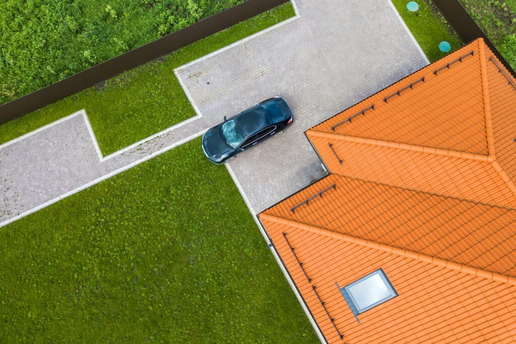 Aerial top view of house shingle roof with attic windows and black car on paved yard with green